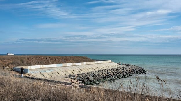 A newly constructed seawall along a coastline, designed to protect coastal infrastructure from rising sea levels and storm surges. The seawall is made of concrete and reinforced with steel, showing a solid and robust barrier. In the background, there are buildings and roads, indicating that the seawall is protecting an urban area.