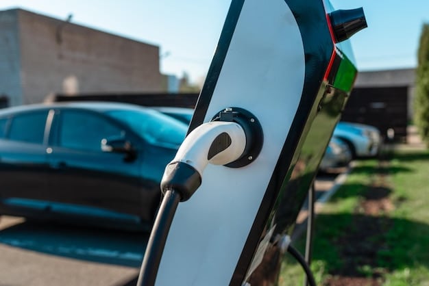 A close-up shot of an EV charging cable plugged into a charging station, with a blurred background showing a row of parked electric cars. The setting is a public charging station with a focus on the charging port and cable.